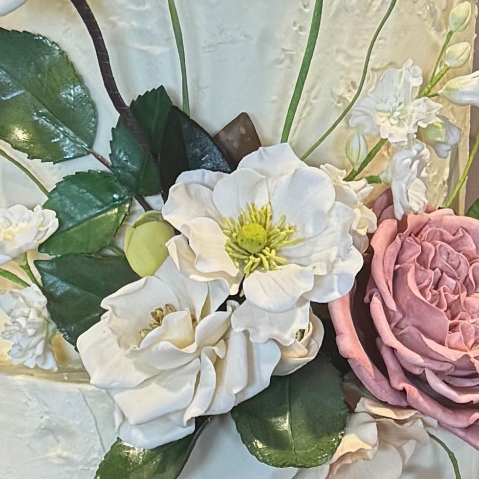 Three-tiered wedding cake with flowers on a wooden stand in a room with a window and decorative wall.
