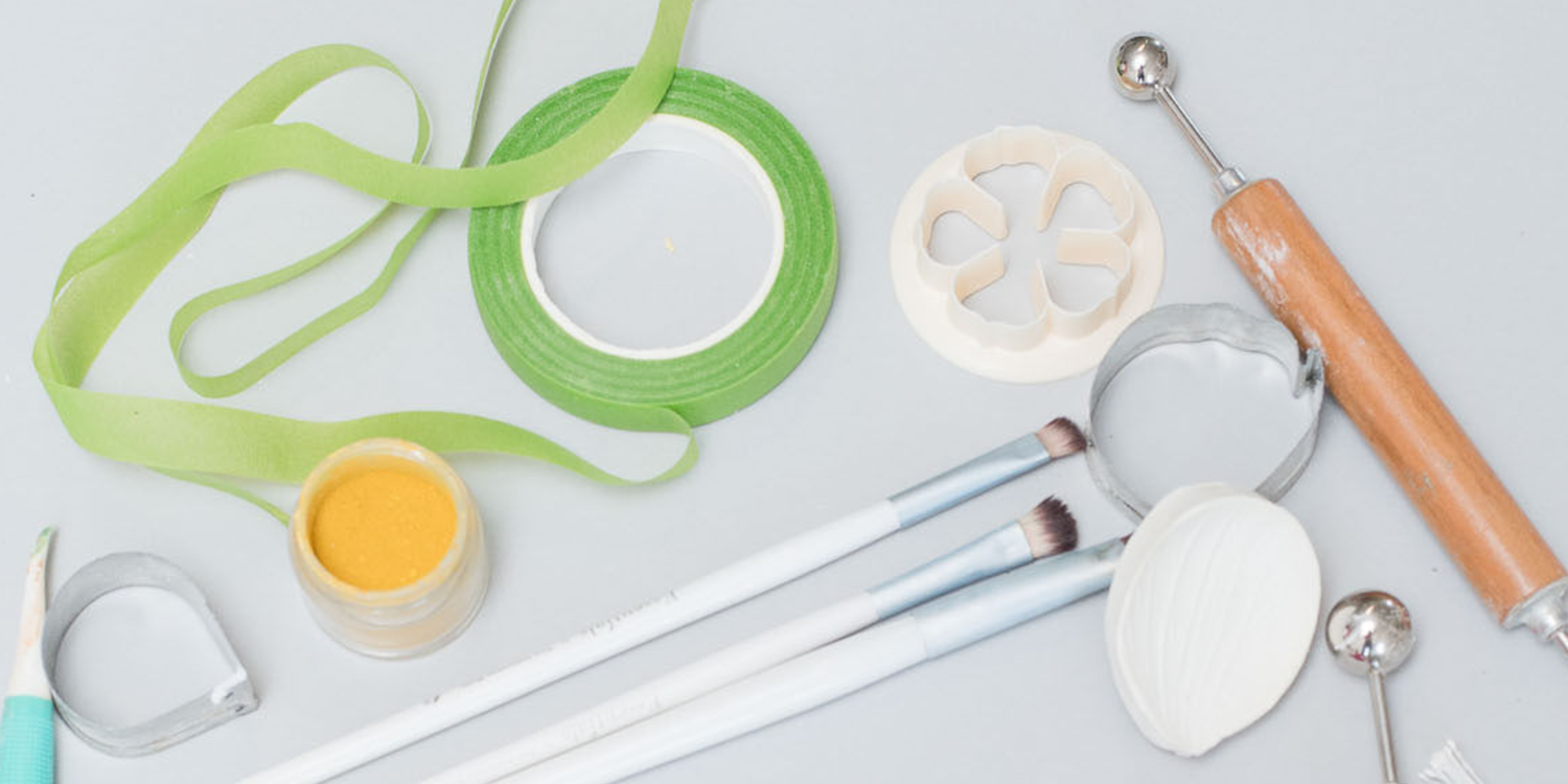 Floral tape, yellow powdered food coloring, paint brushes, steel flower petal cutters, silicone veiners, and wooden ball tools laying on a gray background.